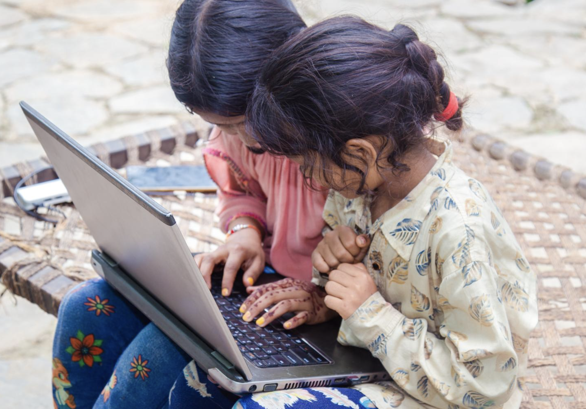 Two girls sitting on the pavement, looking at a computer.