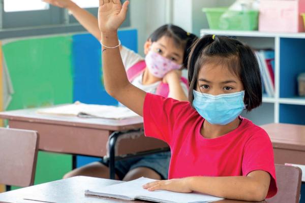 Two children raising hands in classroom, wearing facemasks