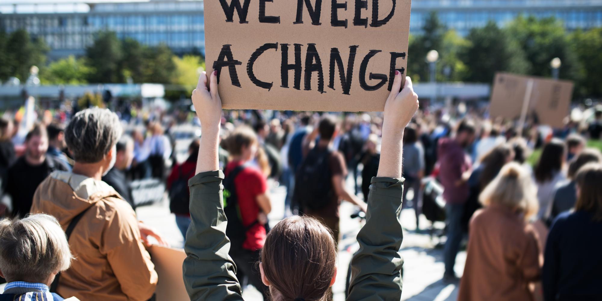 Protestor stands with her back to camera, holding sign that reads "We Need a Change" at a climate change protest